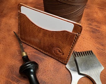 May include: A brown leather card holder with a white card inside. The card holder has a stitched edge and a small logo on the front. The card holder is on a brown leather surface with a leather stitching tool and a awl.