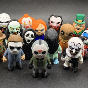 May include: A collection of horror movie character figurines, including Freddy Krueger, Michael Myers, and Jason Voorhees. The figures are brightly colored and detailed, with a variety of costumes and expressions. The figurines are standing on a dark surface.