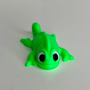 May include: A bright green 3D printed chameleon figurine with black eyes. The chameleon is lying on its belly with its legs outstretched.
