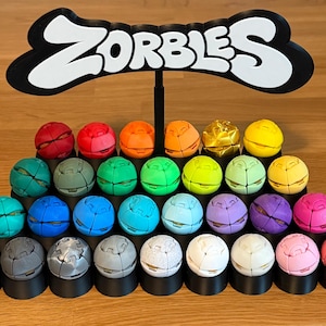 May include: A black display stand featuring the word ZORBLES in white lettering, holding twenty-eight spherical toys in various solid colors including red, orange, yellow, green, blue, purple, pink, white, gray, and black, arranged in rows.