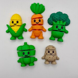 May include: A collection of five colorful vegetable-shaped figurines. The set includes a yellow corn, an orange carrot, a green broccoli, a green bell pepper, and a tan potato. Each figure has a smiling face and arms and legs.