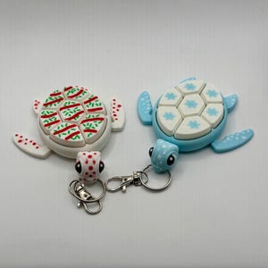 Peut inclure: Deux porte-clés en forme de tortue. L'un est blanc avec des accents rouges et verts, l'autre est bleu clair avec des accents blancs et bleus. Les deux ont des anneaux porte-clés argentés et des mousquetons. Les tortues ont des motifs détaillés sur leurs carapaces et leurs têtes.