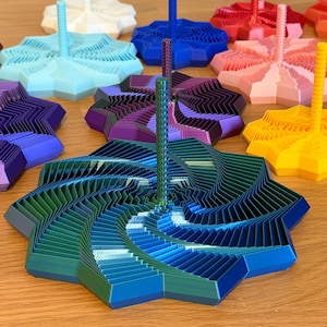 May include: A selection of multi-coloured, geometric, 3D-printed objects. Each has a star-shaped base with layered, angled segments and a central, vertical post. Colours include blue, green, purple, pink, and yellow. The objects are displayed on a wooden surface.