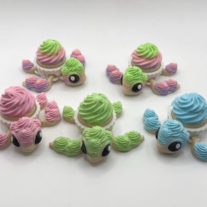 May include: A collection of whimsical turtle-shaped figurines, each adorned with a cupcake-like shell in various pastel colors. The turtles have detailed faces and decorative flipper-like appendages, creating a playful and unique aesthetic.