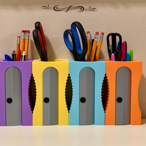 May include: A set of five colourful pencil sharpeners with a grey sharpening mechanism. Each sharpener has a different colour: purple, yellow, blue, light blue, and orange. The sharpeners are filled with various pencils, scissors, and other writing utensils.