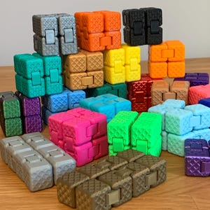 May include: A collection of colorful fidget cubes in various shades, including gray, orange, black, yellow, green, blue, pink, and brown. Each cube is composed of smaller, textured blocks connected by hinges, creating a tactile and engaging toy.