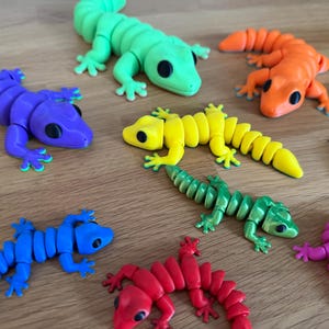 May include: A collection of articulated lizard figurines in various solid colors including purple, lime green, orange, yellow, blue, red, pink, and a metallic green. Each lizard features a segmented body, four legs, and large black circular eyes.