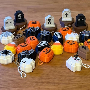 May include: A collection of small Halloween-themed plastic trinkets including orange jack-o'-lanterns, white ghosts, black bats, candy corn, brown coffins, and gray tombstones with R.I.P. or cross symbols, some featuring attached ball chains.