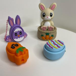 May include: Four colorful Easter-themed figurines: a purple bunny with a white face, an orange carrot with green leaves, a tan bunny sitting in a brown basket, and a blue egg with pink and yellow stripes.
