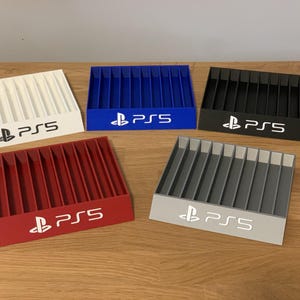 May include: Five different colored plastic storage boxes for video game discs. Each box has a PlayStation 5 logo and is labeled "PS5". The boxes are white, blue, red, gray, and black.