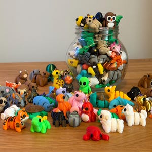 May include: A selection of colourful animal figurines, including a tiger, owl, and alien, are scattered on a wooden surface. Many more figurines are inside a clear glass jar. The figurines are made of clay or a similar material.