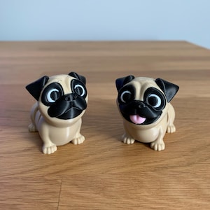 May include: Two tan pug figurines with black ears, noses, and eye surrounds. The pugs have large, expressive eyes and are posed in a seated position. One pug has its tongue sticking out. The figurines are on a wooden surface.