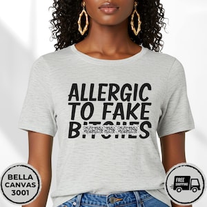 May include: Light gray Bella Canvas 3001 t-shirt with the text "ALLERGIC TO FAKE B****S" in black. The word "B****S" is partially obscured. The shirt is short-sleeved and has a crew neck.