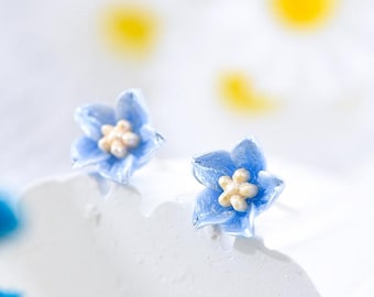 May include: A pair of blue flower stud earrings with white centers. The earrings are on a white surface.