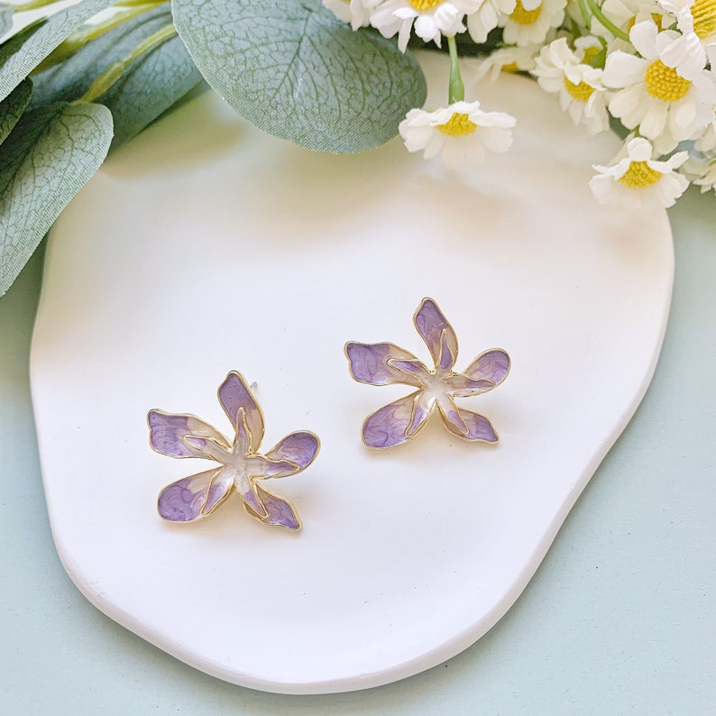 May include: A pair of purple and gold flower-shaped earrings. The earrings are on a white surface.