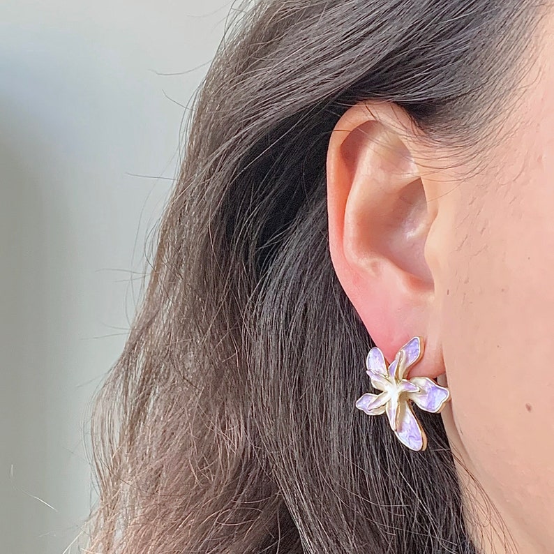 May include: A pair of purple flower-shaped earrings with a gold setting. The earrings are worn by a person with long brown hair.