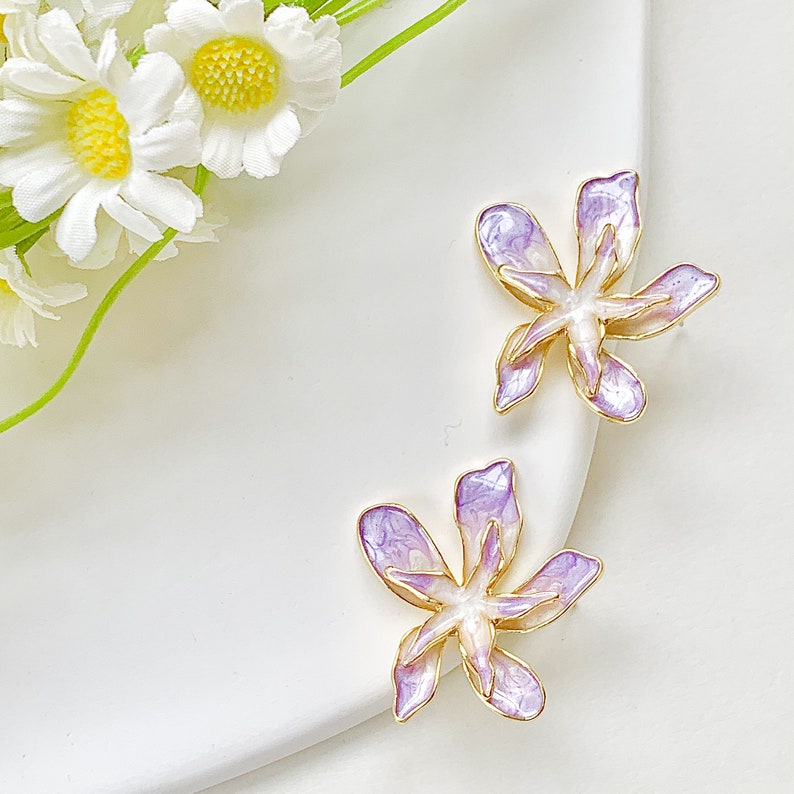 May include: A pair of purple and white flower-shaped earrings with gold accents. The earrings are delicate and feminine.