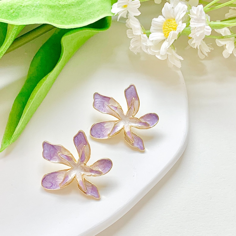 May include: A pair of purple and white flower-shaped earrings with gold accents. The earrings are on a white surface.