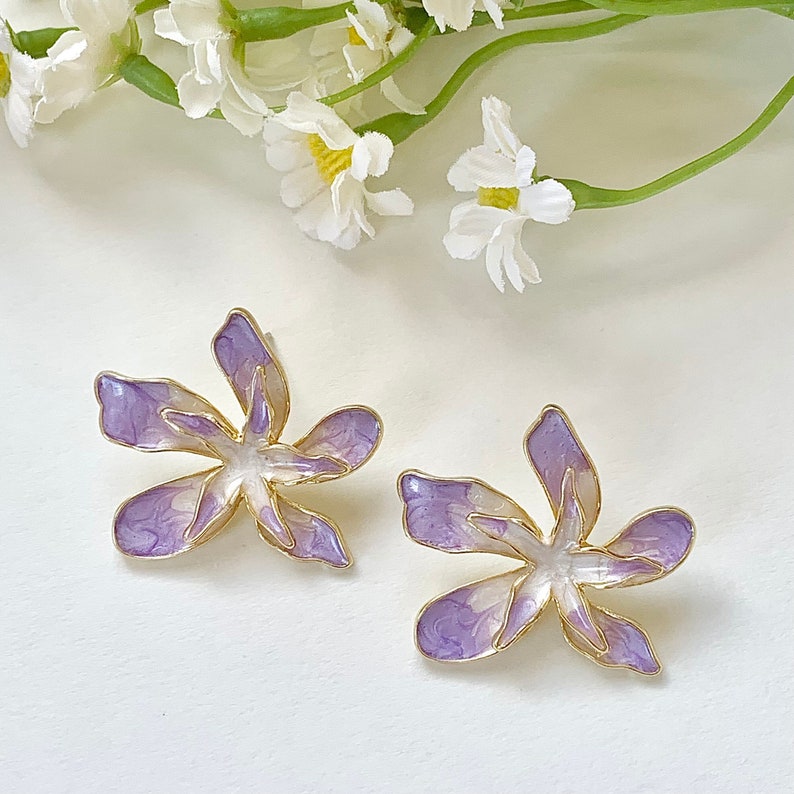 May include: A pair of purple and white flower-shaped earrings with gold accents. The earrings are made of metal and have a delicate, feminine design.