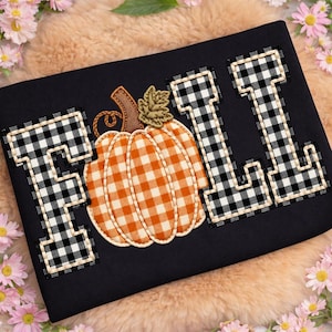May include: A black fabric item featuring an embroidered applique design. The word FALL is spelled out with letters in a black and white buffalo check pattern. The letter A is replaced by an orange and white gingham pumpkin with a brown stem.