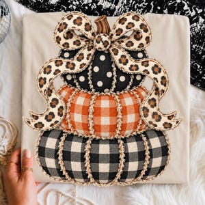 May include: A beige t-shirt featuring a stacked pumpkin graphic. The top pumpkin is black with white polka dots, the middle is orange plaid, and the bottom is black and white buffalo check. A large leopard print bow sits on top.