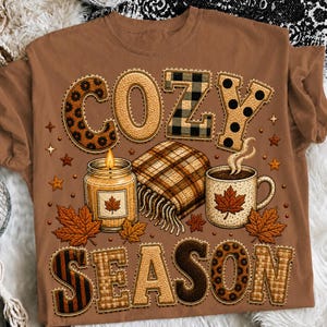 Op de afbeelding: Een bruin t-shirt met de tekst COZY SEASON in blokletters met patronen. De afbeelding bevat een brandende kaars in een pot, een gevouwen geruite deken met franjes en een dampende mok, versierd met oranje esdoornbladillustraties.