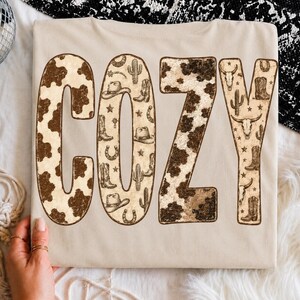 Puede incluir: Una camiseta beige con la palabra COZY en letras grandes. Las letras C y Z tienen un patrón de manchas de vaca marrón y blanco, mientras que las letras O y Y presentan un fondo beige con pequeñas ilustraciones marrones de botas vaqueras, sombreros, cactus, herraduras y cráneos.