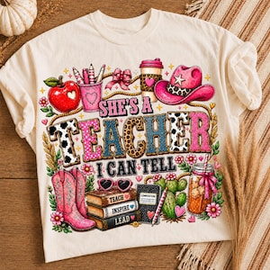 May include: A cream-colored t-shirt featuring a colorful graphic print with the text 'SHE'S A TEACHER I CAN TELL'. The design includes a red apple, pink cowboy hat, pink boots, a stack of books labeled 'TEACH', 'INSPIRE', 'LEAD', and a cactus.