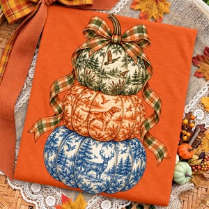 Pode incluir: Uma t-shirt laranja com um gráfico de três abóboras empilhadas atadas com uma fita xadrez. A abóbora superior mostra um lago com patos, a do meio um cão numa floresta e a inferior um veado numa paisagem florestal com neve.