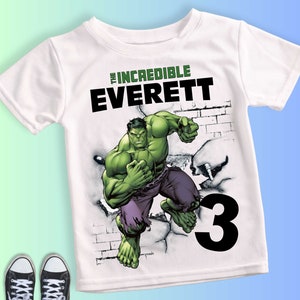 May include: White t-shirt featuring a graphic of the Hulk breaking through a wall. The shirt includes the text "THE INCREDIBLE EVERETT" in green and black, and the number "3" in black. Black and white sneakers are visible at the bottom.
