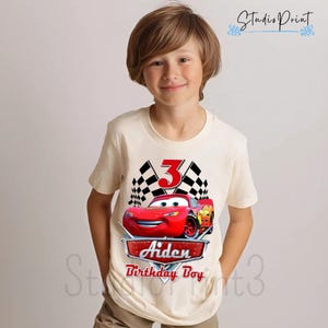 Puede incluir: Una camiseta blanca con un gr&aacute;fico de coche de carreras rojo, banderas a cuadros y el n&uacute;mero 3. El texto "Aiden Birthday Boy" est&aacute; impreso debajo del coche.
