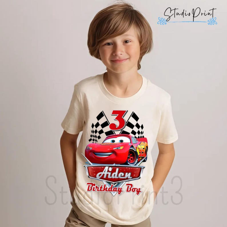 Puede incluir: Una camiseta blanca con un gr&aacute;fico de coche de carreras rojo, banderas a cuadros y el n&uacute;mero 3. El texto "Aiden Birthday Boy" est&aacute; impreso debajo del coche.