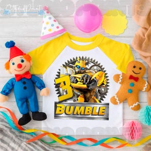 Puede incluir: Una camiseta raglán blanca y amarilla con un gráfico de Bumblebee, el número 3 y la palabra "BUMBLE". La imagen incluye un muñeco de payaso, un hombre de jengibre, decoraciones de fiesta y un gorro de fiesta.