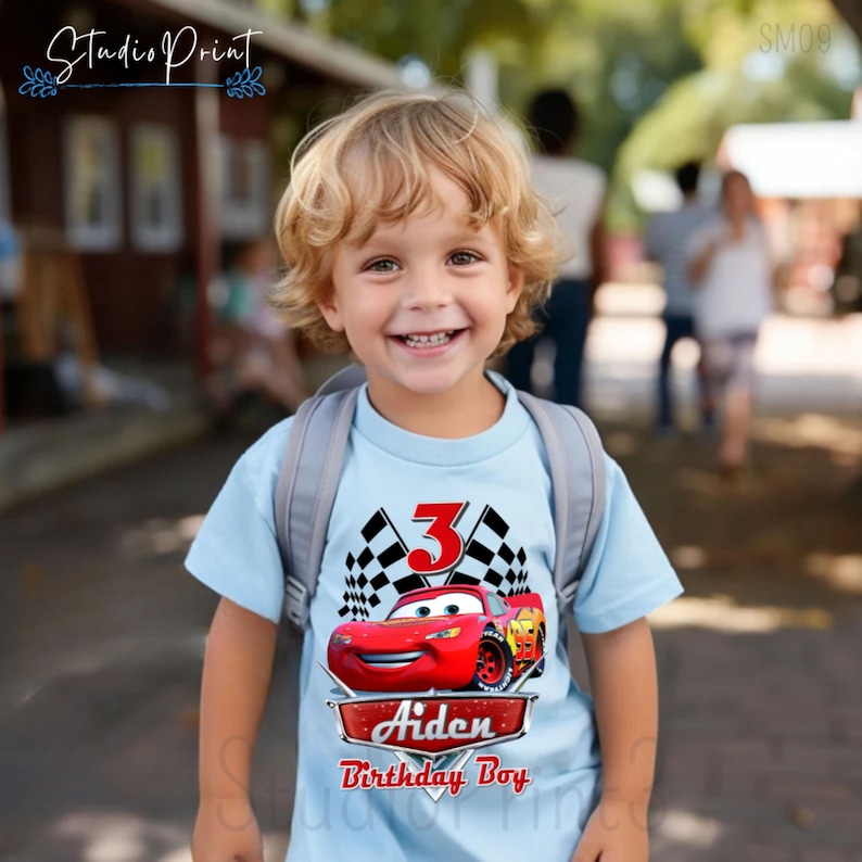 Puede incluir: Una camiseta azul claro con un gr&aacute;fico de un coche de carreras rojo, banderas a cuadros y el n&uacute;mero 3. El texto "Aiden Birthday Boy" est&aacute; impreso debajo del coche.