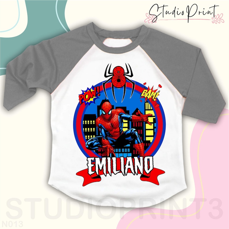 May include: A white and gray baseball t-shirt with a red and blue Spiderman graphic. The graphic includes the words "POW!" and "BAM!" and the name "EMILIANO".