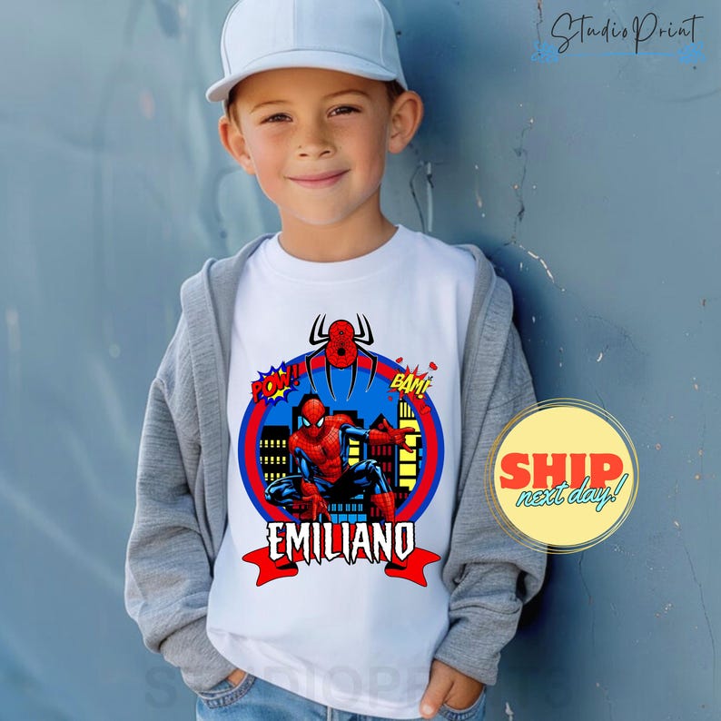May include: A white t-shirt with a blue, red, and yellow graphic of Spiderman in a city scene. The graphic includes the words 'POW!' and 'BAM!' and the name 'EMILIANO'.