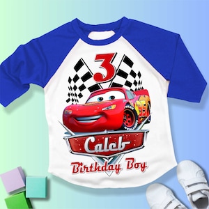 May include: A white and blue baseball t-shirt with a red race car and checkered flags. The number 3 is on the shirt. The text "Caleb Birthday Boy" is on the shirt.