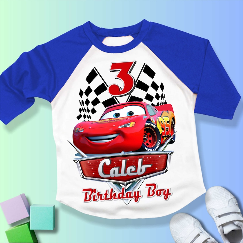 May include: A white and blue baseball t-shirt with a red race car and checkered flags. The number 3 is on the shirt. The text "Caleb Birthday Boy" is on the shirt.