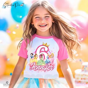 May include: A white and pink raglan t-shirt with a princess design. The shirt displays the number 6 with a crown, princess illustrations, and the name "Jennifer". The t-shirt is worn by a young girl.
