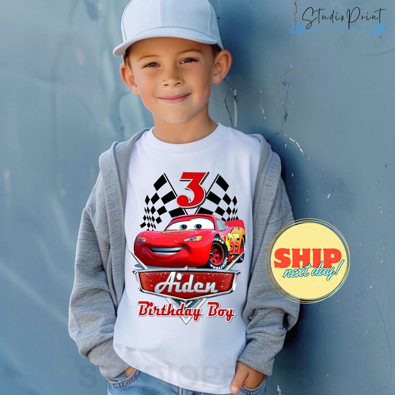 Puede incluir: Camiseta blanca con un coche de carreras rojo, banderas a cuadros y el n&uacute;mero 3. El texto "Aiden Birthday Boy" est&aacute; impreso debajo del coche.