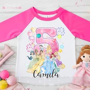 May include: A white and pink raglan-style shirt with a large number 5 graphic, featuring Disney princesses and the name "Camila". The number is decorated with balloons and fireworks. The sleeves are bright pink.