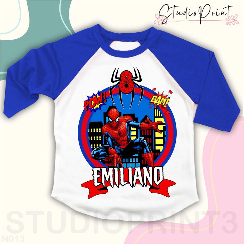 May include: A white and blue baseball t-shirt with a red and blue Spiderman graphic. The graphic includes the words "POW!" and "BAM!" and the name "EMILIANO".