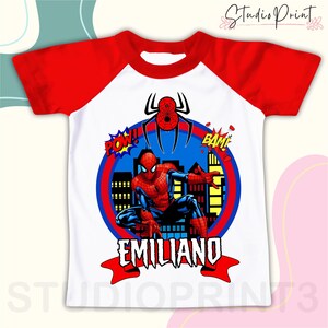 May include: White and red raglan t-shirt with a Spiderman graphic. The graphic features Spiderman in a city scene with the words "POW!" and "BAM!" and the name "EMILIANO" below.