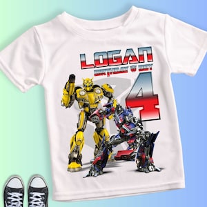 Könnte beinhalten: Weißes T-Shirt mit einem Aufdruck von Bumblebee und Optimus Prime aus Transformers. Auf dem Shirt stehen der Name "LOGAN", "BIRTHDAY BOY" und die Zahl "4". Schwarze und weiße Turnschuhe sind unten im Bild zu sehen.