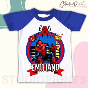 May include: A white and blue raglan t-shirt with a red and blue Spiderman graphic, a red and blue cityscape, and the text "POW!" and "BAM!" in red. The name "EMILIANO" is written in red on a red and white ribbon.
