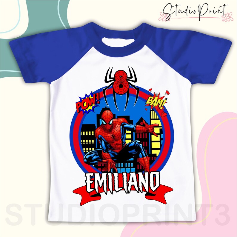 May include: A white and blue raglan t-shirt with a red and blue Spiderman graphic, a red and blue cityscape, and the text "POW!" and "BAM!" in red. The name "EMILIANO" is written in red on a red and white ribbon.