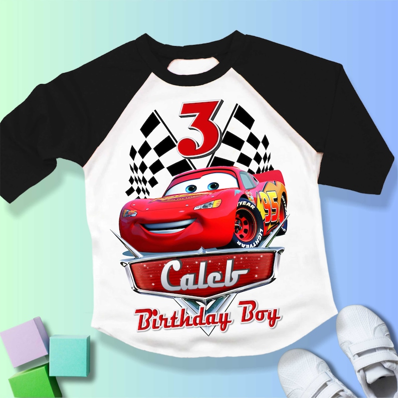May include: A white and black baseball style t-shirt with a red race car and checkered flags. The number 3 is in red and the text "Caleb Birthday Boy" is in silver.
