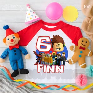 以下が含まれることがあります： 「6」のグラフィックと「Finn」という名前が入った白と赤のラグランシャツ。シャツはRobloxのキャラクターデザインです。画像には、ピエロの人形、ジンジャーブレッドマン、パーティーの飾りも含まれています。