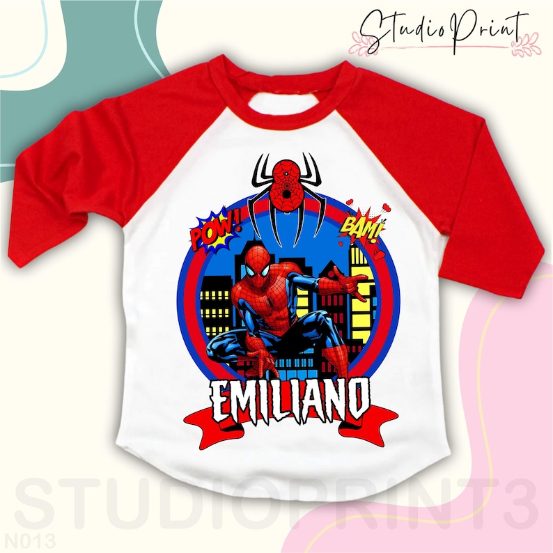 May include: Red and white baseball style t-shirt with a Spiderman graphic, city skyline, and the words "POW!" and "BAM!"  The shirt also has the name "EMILIANO" in a red banner.