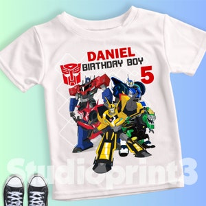 Könnte beinhalten: Weißes T-Shirt mit einer farbenfrohen Grafik von Transformers-Charakteren. Der Text "DANIEL BIRTHDAY BOY 5" ist rot. Das Transformers-Logo ist ebenfalls zu sehen.